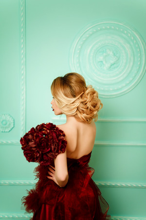 Sexy stylish blonde beautiful woman near green decorated wall with bouquet of red camellia flowersの写真素材