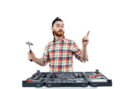 Redhead Bearded man wearing a baseball cap and shirt holding a Claw hammer from a toolbox in front of him. Isolated on white.の写真素材