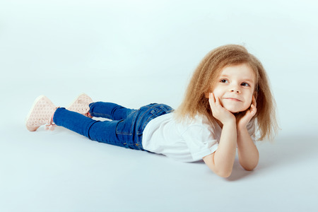 little girl 4 years old with curly hair wearing white shirt, blue jeans lying on the floor, smiling and looking at camera, hands holding her headの写真素材