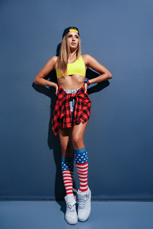 glamor sexy smiling beautiful young woman model in sport shirt and hat and Red checkered shirt, red wristlet watch, white sneakers, leggings with USA flag, leaned on textured wallの写真素材