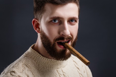 Portrait of handsome serious bearded man in white sweater holding cigar on black background