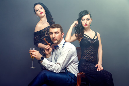 Portrait of handsome man with drink and cigar sitting on chair surrounded by two beautiful black-haired women in dresses.Smoky background.Studio shotの写真素材