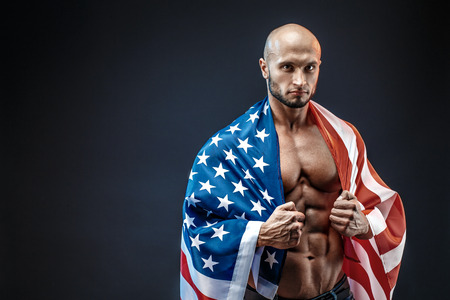 Sexy shredded man with American flag on shoulder looking at camera. Strong bodybuilder with six pack, perfect abs, shoulders, biceps, triceps and chest, personal fitness trainer flexing his muscles.の写真素材