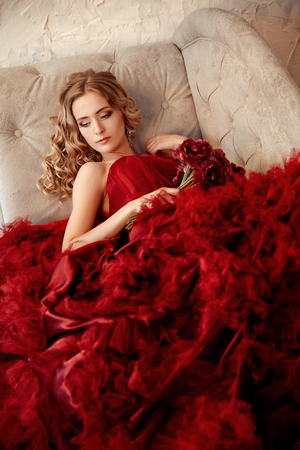 Sexy stylish blonde beautiful woman on the sofa. bouquet of flowers, peonies, pompons. Red dress.の写真素材