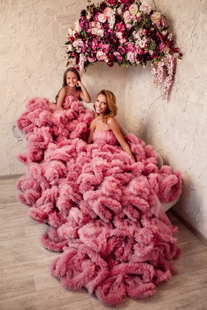 Beautiful slim blonde mom in dress and young daughter with flowers on a background. Studio. Interior.の写真素材