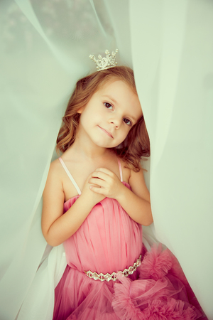 Portrait of cute little girl with hands on chest in pink dress and diadem looking at cameraの写真素材