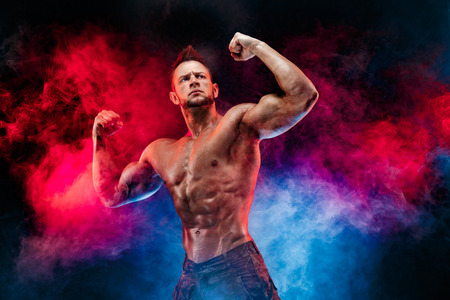 Close up on perfect abs. Strong bodybuilder with six pack. Man with perfect abs, shoulders,biceps, triceps and chest, personal fitness trainer flexing his muscles in blue, red smokeの写真素材