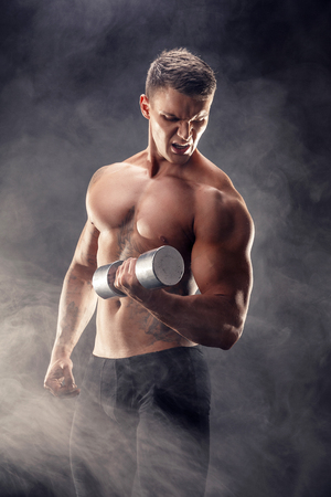 Handsome bodybuilder doing exercise on biceps with dumbbell. Studio shot. Smoke.の写真素材