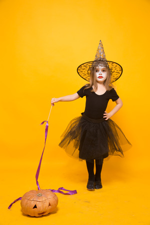 Small girl in Halloween witch costume on groom with pumpkin aside isolated on orange background.の写真素材