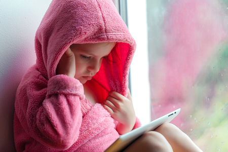 Cute little girl in pink bathrobe using tablet pc. Sitting on the window sill.の写真素材