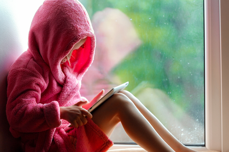 Cute little girl in pink bathrobe using tablet pc. Sitting on the window sill.の写真素材