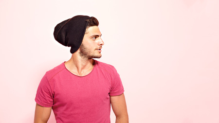 Good-looking brunette guy standing dressed in pink t-shirt and brown hat looking away.
