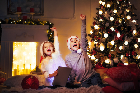 Excited boy and girl sitting near Christmas tree, holding their hands up and looking at camera.の写真素材
