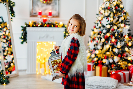 Cute little girl in checkered dress standing in brightly decorated room and holding Christmas decoration.の写真素材