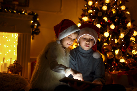 Excited boy and girl sitting near Christmas tree, playing tablet game together.の写真素材