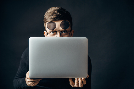 Young smart man wearing flip-up glasses covering face with laptop looking at camera.の写真素材
