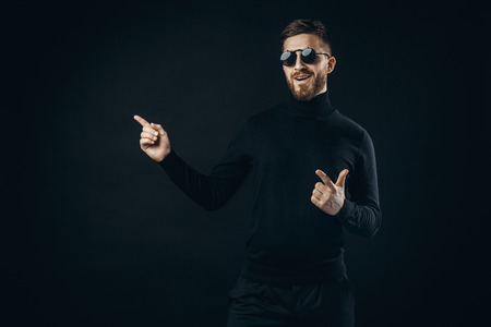 Handsome bearded man in black and flip-up sunglasses smiling and pointing away on black.