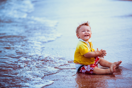 Little kid, boy in yellow shirt, red shorts, sitting on the beach close to  water, playing laughing, smilingの写真素材
