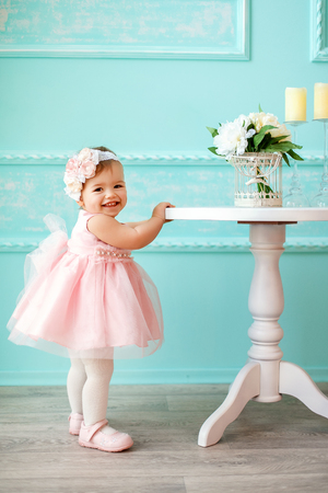 Portrait of a beautiful little baby in pink dress standing and holding table with flowers.の写真素材