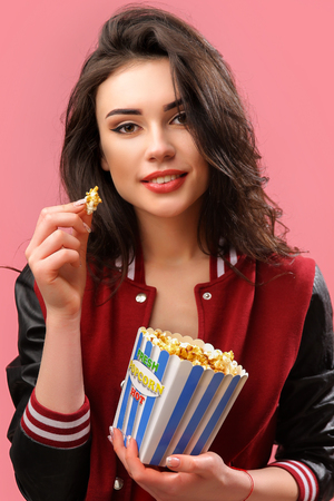Alluring young brunette in popcorn bag looking playfully at camera having bite on pink background.の写真素材