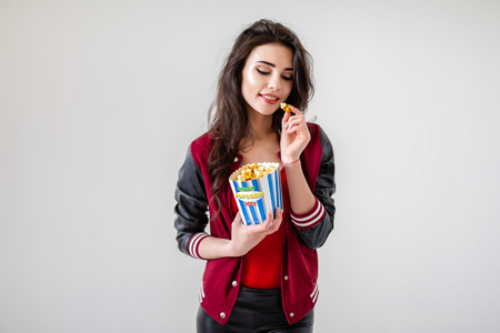 Alluring young brunette in popcorn bag looking playfully at camera having bite on white or gray background.の写真素材