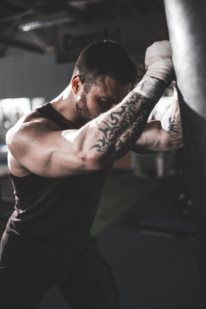 Bearded Male boxer training with punching bag in dark sports hall. Young tattoed boxer training on punching bag. Male boxer as exercise for the big fight. Boxer hits punching bag.の写真素材