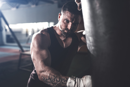 Bearded Male boxer training with punching bag in dark sports hall. Young tattoed boxer training on punching bag. Male boxer as exercise for the big fight. Cool tone.の写真素材