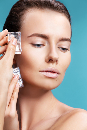 Closeup portrait of beautiful young woman applies the ice to face on a blue background. Skin care concept.の写真素材