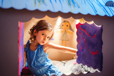 Content pretty little girl sitting inside of colored carton playhouse looking at camera through small window.の写真素材