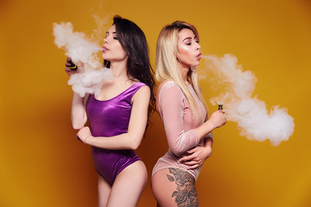 Two young women in bodysuits puffing vapor on yellow background in studio.