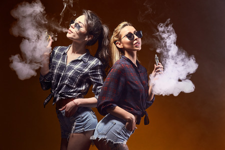 Side view of trendy young female in glasses and shorts standing and smoking on studio backgroundの写真素材