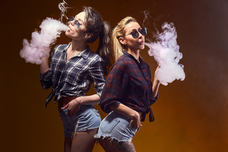 Side view of trendy young female in glasses and shorts standing and smoking on studio backgroundの写真素材