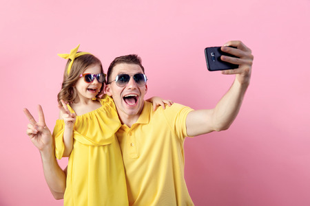 Portrait of happy family of two people in yellow. Daddy and little daughter doing selfie with smartphone on pink background. Horizontal color photography.の写真素材