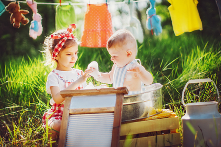 Little cute brother in apron bathing in pelvis and pretty sister in checkered dress headband laughing sitting together outdoorsの写真素材
