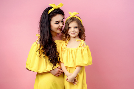 Crop view of positive brunette mother holding smilling daughter in same yellow dress looking at camera in studio on pink backgroundの写真素材