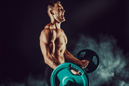 Athletic man doing exercises with heavy barbell at biceps. Photo of strong male with naked torso on dark background with smoke. Strength and motivation.の写真素材