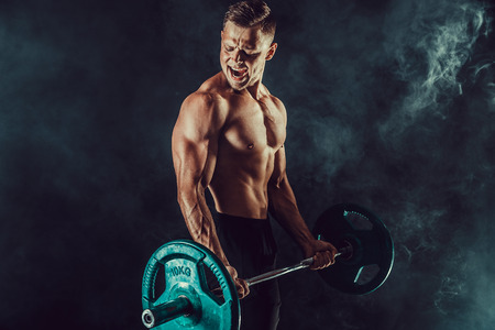 Athletic man doing exercises with heavy barbell at biceps. Photo of strong male with naked torso on dark background with smoke. Strength and motivation.の写真素材