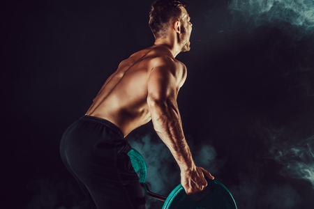 Strong athletic man pumping up muscles workout bodybuilding. muscular bodybuilder handsome men doing exercise for muscles of back with a barbell on dark background with smoke. Strength and motivation.の写真素材