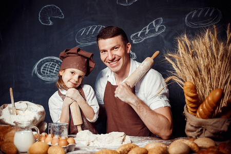 Happy adult man with little girl wearing aprons and holding rolling pins looking at camera while making bread doughの写真素材