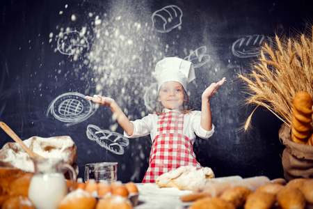 Adorable little girl in checkered apron and hat throwing flour powder in air with eyes closed having fun while making doughの写真素材