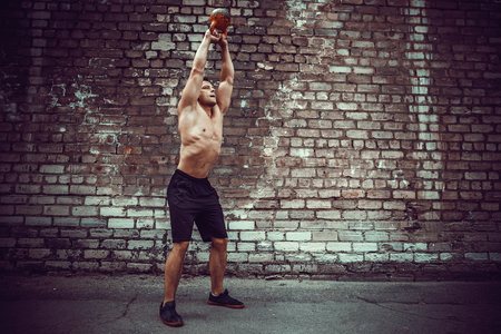 Athletic man working out with a kettlebell in front of brick wall. Strength and motivation. Outdoor workout.の写真素材