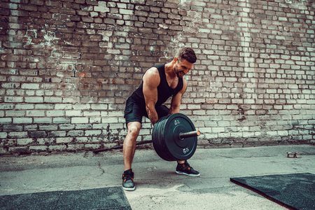 Athletic man working out with a barbell in front of brick wall. Strength and motivation. Outdoor workout. Exercise for the muscles of the backの写真素材