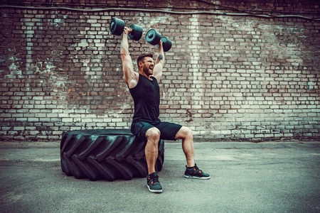 Athletic man working out with a dumbbell in front of brick wall. Strength and motivation. Outdoor workout. Exercise for the shoulder muscles, deltoid.の写真素材