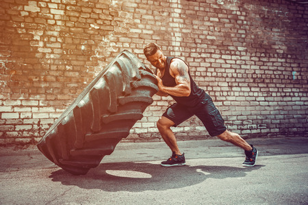 Muscular bearded tattooed fitness man moving large tire in street gym. Concept lifting, workout training.の写真素材