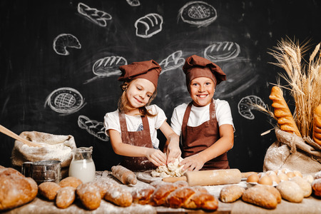 Adorable girl with brother in aprons on table with bread loaves making fresh dough and having fun