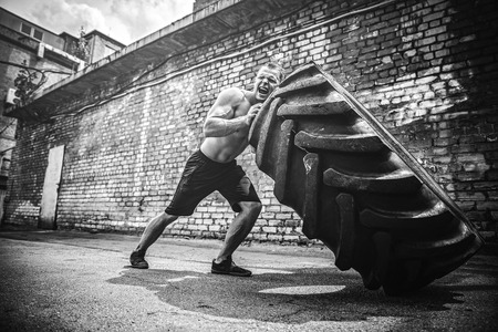 Muscular bearded tattooed fitness shirtless man moving large tire in street gym. Concept lifting, workout training.の写真素材
