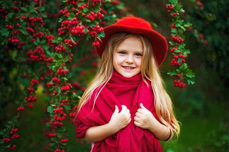 little blonde girl in a red hat, scarf in a park near a treeの写真素材