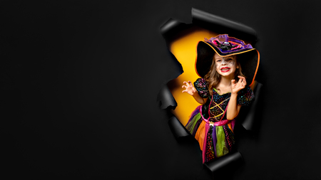 Happy Halloween. Laughing funny child girl in a witch costume of halloween looking, smiling and scares through a hole of black and yellow paper background. Copyspaceの写真素材