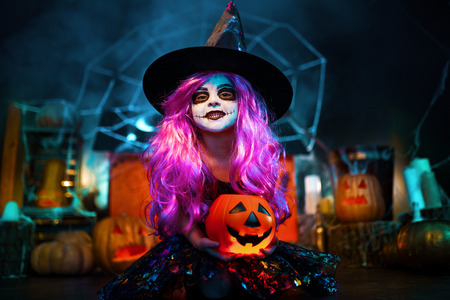 Happy Halloween. A little beautiful girl in a witch costume celebrates at home in an interior with pumpkins and cardboard magic house on the background. Scares at camera.の写真素材