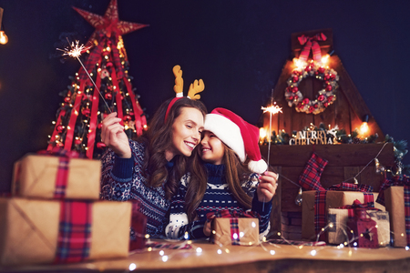 Happy mother in sweater, Christmas deer antlers and little girl in santa helper hat with sparklers in hands, gift boxes on foreground. Having fun over living room, christmas tree, fireplace backgroundの写真素材